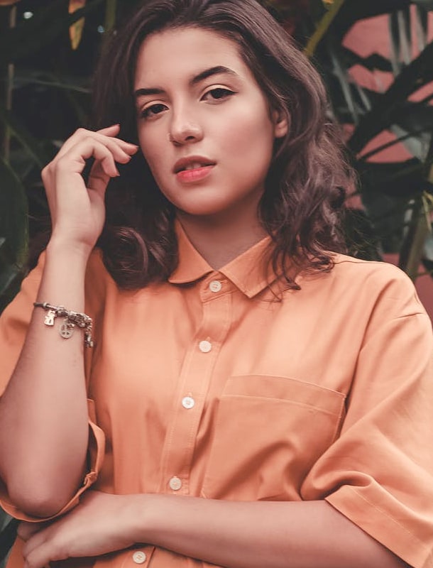 Brown haired lady wear orange shirt, greenery on the background.